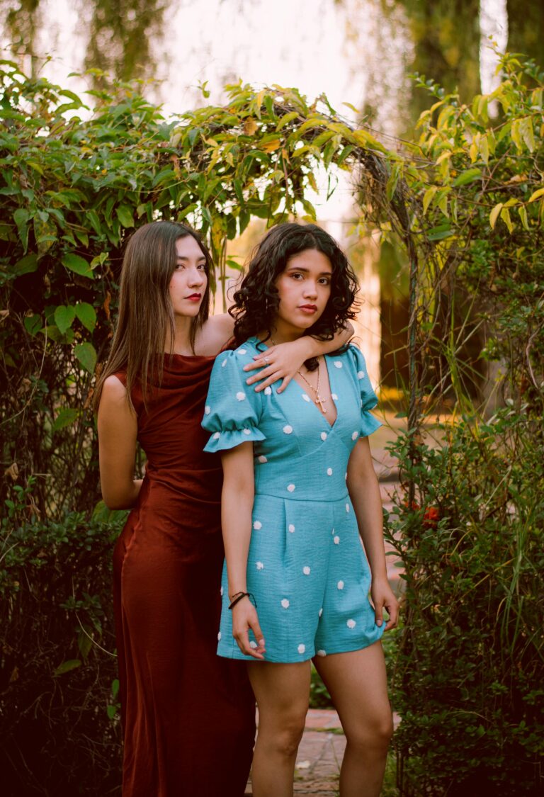 Two young women in stylish outfits posing in a garden setting, surrounded by lush greenery.
