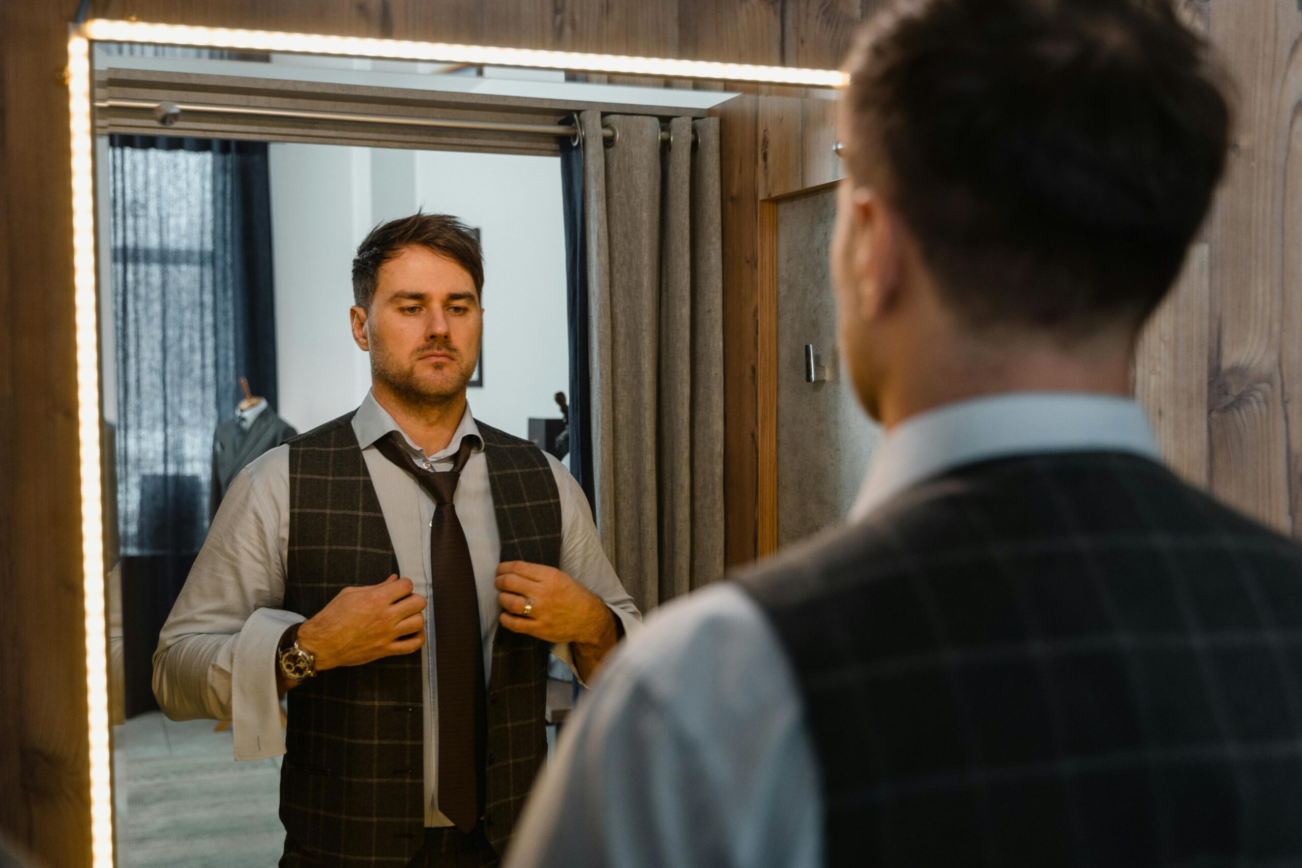 A well-dressed man in a vest adjusts his tie in an elegant indoor setting.