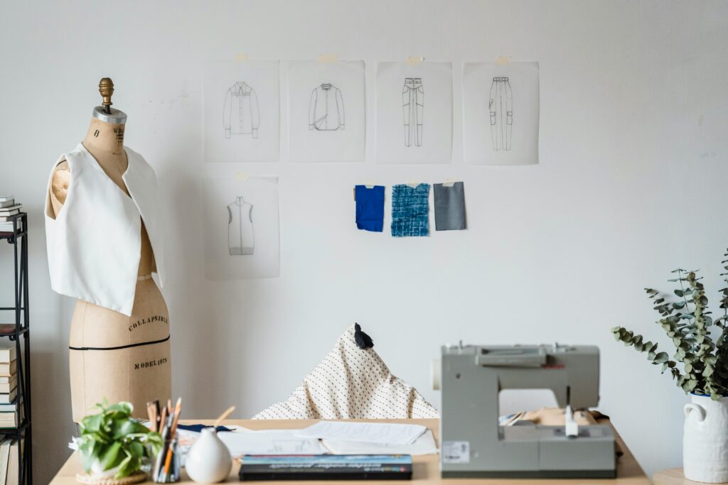 Table with sewing machine placed near mannequin with garment at white wall with drafts of cloth and samples of textile