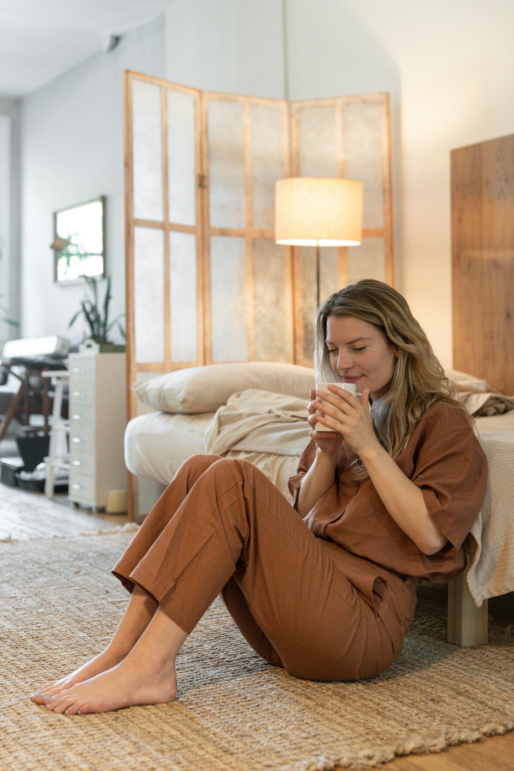 Woman in pajamas enjoying coffee in a cozy, minimalist bedroom setting.